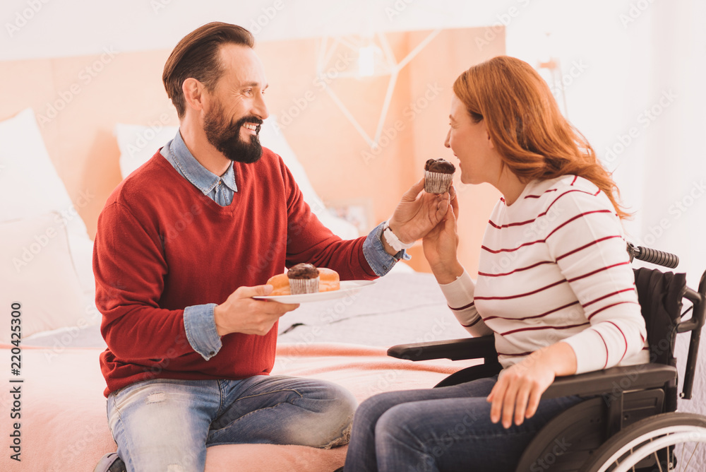 Try it. Cheerful man feeding his disabled woman with a cupcake while resting at home