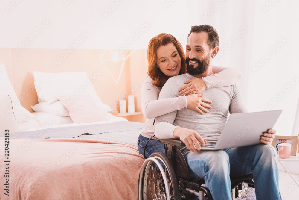Love you so much. Cheerful handsome disabled man using a laptop while his wife embracing him Love you so much. Cheerful handsome disabled man using a laptop while his wife embracing him