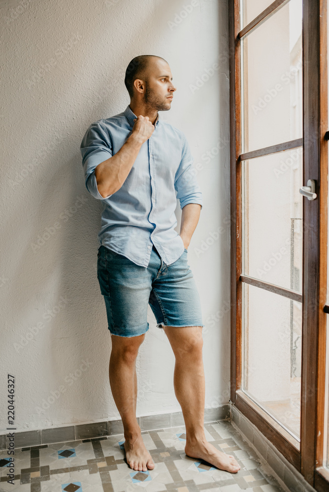 Brutal man in the blue shirt and blue jeans shorts stands on the floor of  the flat with white walls holding his collar near the window in Spain Stock  Photo | Adobe