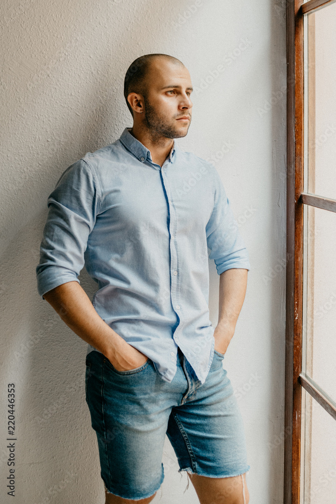Tanned brutal man in the blue shirt and blue jeans shorts thinking holding hands in pockets in the the flat with white walls near the window in Spain