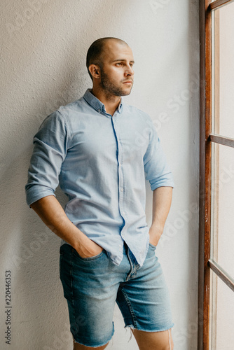 Obraz na płótnie Tanned brutal man in the blue shirt and blue jeans shorts thinking holding hands in pockets in the the flat with white walls near the window in Spain w salonie