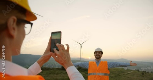 Rear of the female woman, worker of the renewable wind energy station, taking a photo of her male co-worker in the helmet on the smartphone. Huge winmills turbines on the background. photo