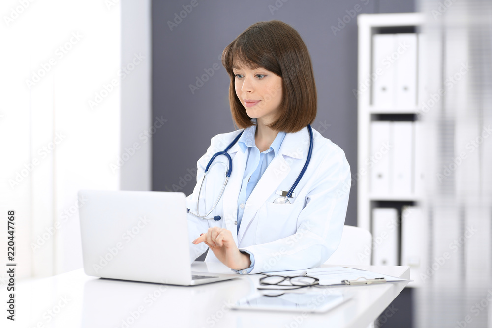 Doctor typing on laptop computer while sitting at the white table in hospital office. Physician at work. Medicine and healthcare concept Doctor typing on laptop computer while sitting at the white table in hospital office. Physician at work. Medicine and healthcare concept