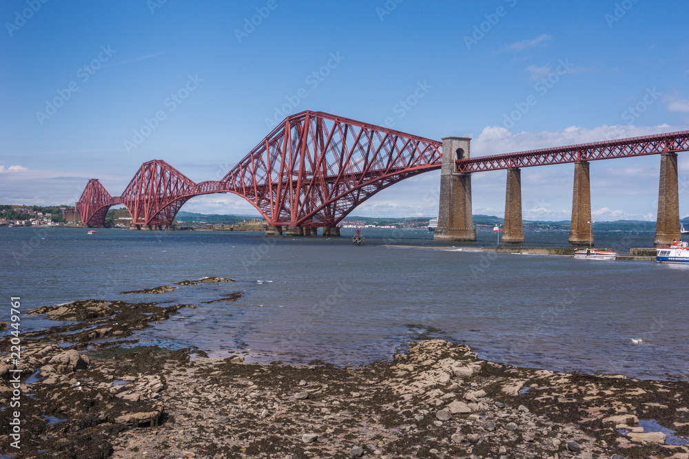 Queensferry, Scotland, UK - June 14, 2012: Closeup of Red metal iconic Forth Bridge for trains over Firth of Forth between blue sky and blue water. Rocky shore up front. Queensferry, Scotland, UK - June 14, 2012: Closeup of Red metal iconic Forth Bridge for trains over Firth of Forth between blue sky and blue water. Rocky shore up front.
