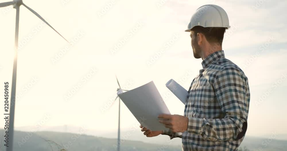 Portrait shot the man in a helmet, environmental and safe power engineer, standing with some documents while watching the work of the big windmill turbine. Portrait shot the man in a helmet, environmental and safe power engineer, standing with some documents while watching the work of the big windmill turbine.
