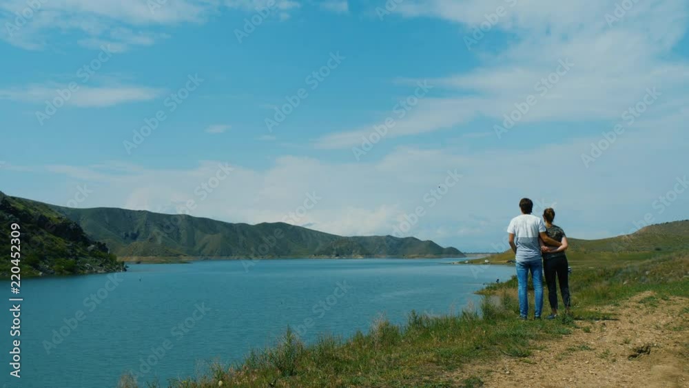 Back view of young couple standing on the shore of lake. Man hugs woman gently Back view of young couple standing on the shore of lake. Man hugs woman gently