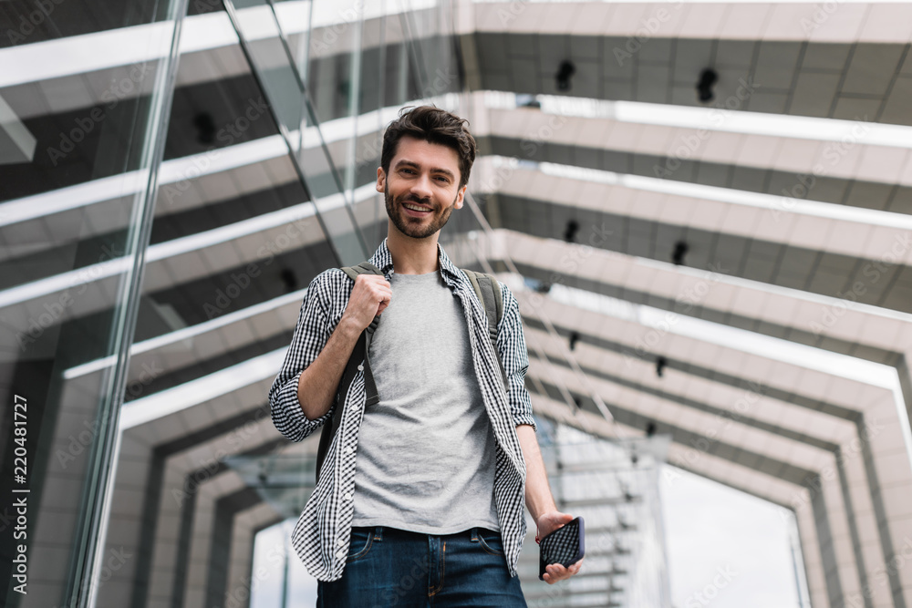 Portrait of happy smiling university students with backpack and smartphone going to take an exam. Bearded hipster wearing stylish casual clothes holding mobile in hand. 