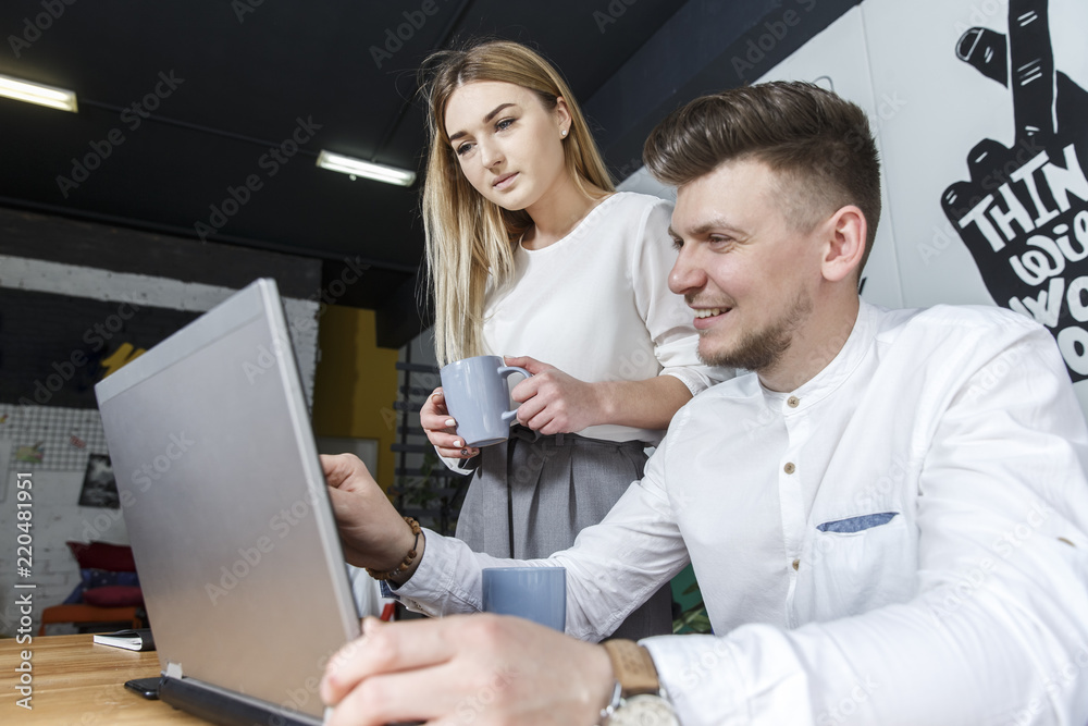 A picture of businessman sitting at table. He is looking at laptop nad holding hand near it. Girl is standing besides man and holds a cup of coffee. She looks at the laptop screen A picture of businessman sitting at table. He is looking at laptop nad holding hand near it. Girl is standing besides man and holds a cup of coffee. She looks at the laptop screen