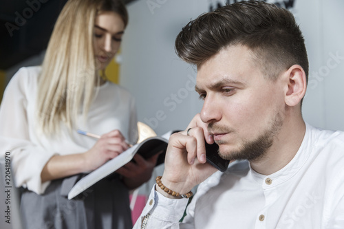 A picture of man looking forward and talking on the phone. He is serious. Girl is standing besides him and looking at journal. She is writing down.