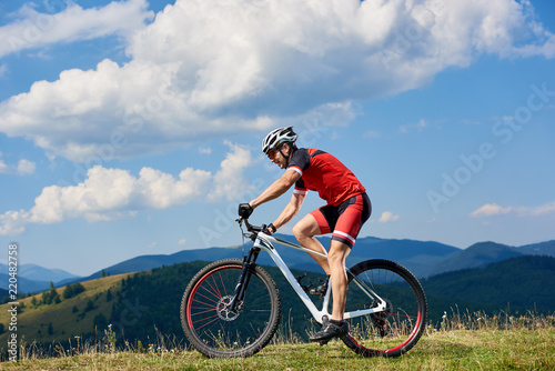 Athlete sportsman bicyclist in professional sportswear and helmet riding cross country bicycle on summer day. Mountains view and cloudy sky on background. Active lifestyle and outdoor sport concept © anatoliy_gleb