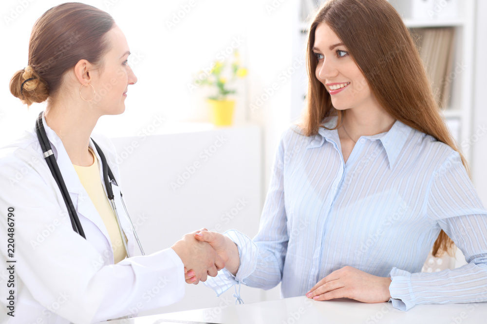 Doctor and patient shaking hands while sitting at the desk near window Doctor and patient shaking hands while sitting at the desk near window