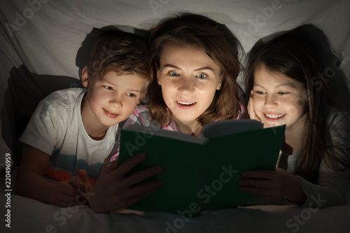 Mother reading bedtime story to her little children under blanket in evening