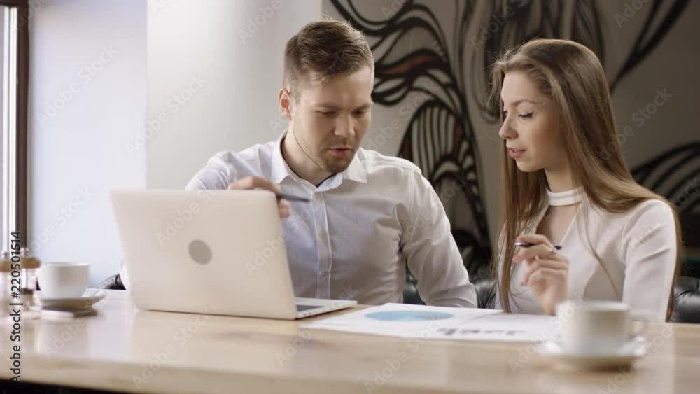 Two young people work with a laptop in a cafe. They are discussing and preparing the contract. It is a business meeting. Man and woman are sitting in the cafe at the table during break. Couple are Two young people work with a laptop in a cafe. They are discussing and preparing the contract. It is a business meeting. Man and woman are sitting in the cafe at the table during break. Couple are