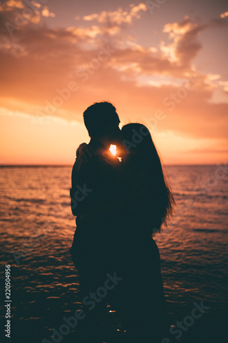 Giovane coppia di ragazzo e ragazza si baciano abbracciandosi di fronte al tramonto rosso fuoco in riva al mare sulla spiaggia. Vista controluce, silhouette.  photo