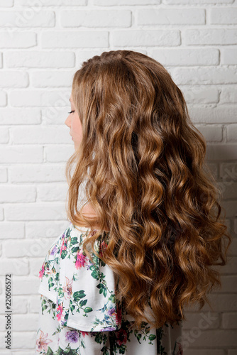 Hairstyle long curls on the head of a brown-haired woman at the back close-up against a white brick wall.