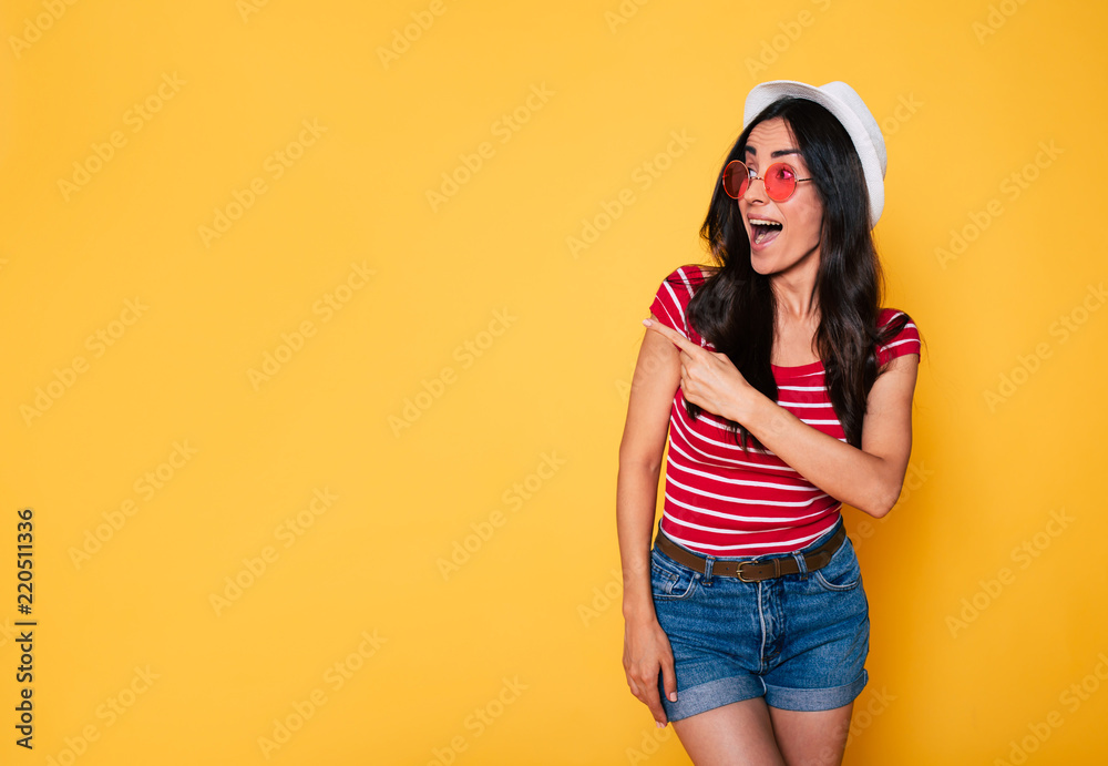 Cute girl pointing away. Happy beautiful young hipster woman in hat and sunglasses posing over yellow background Cute girl pointing away. Happy beautiful young hipster woman in hat and sunglasses posing over yellow background