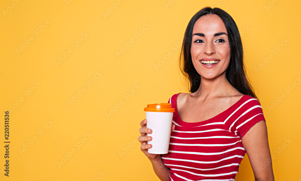 Smiling attractive woman in casual clothes with coffee cup in hands posing over yellow background