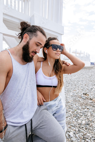 Young couple of fit caucasian woman and bearded hipster man with hair knot having rest after outdoor workout and listen to music through headphones on the lake shore. photo