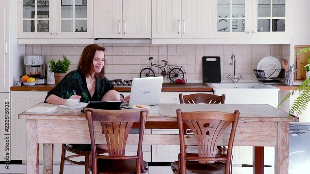 Young woman in bathrobe working on laptop and doing notes by table in kitchen
