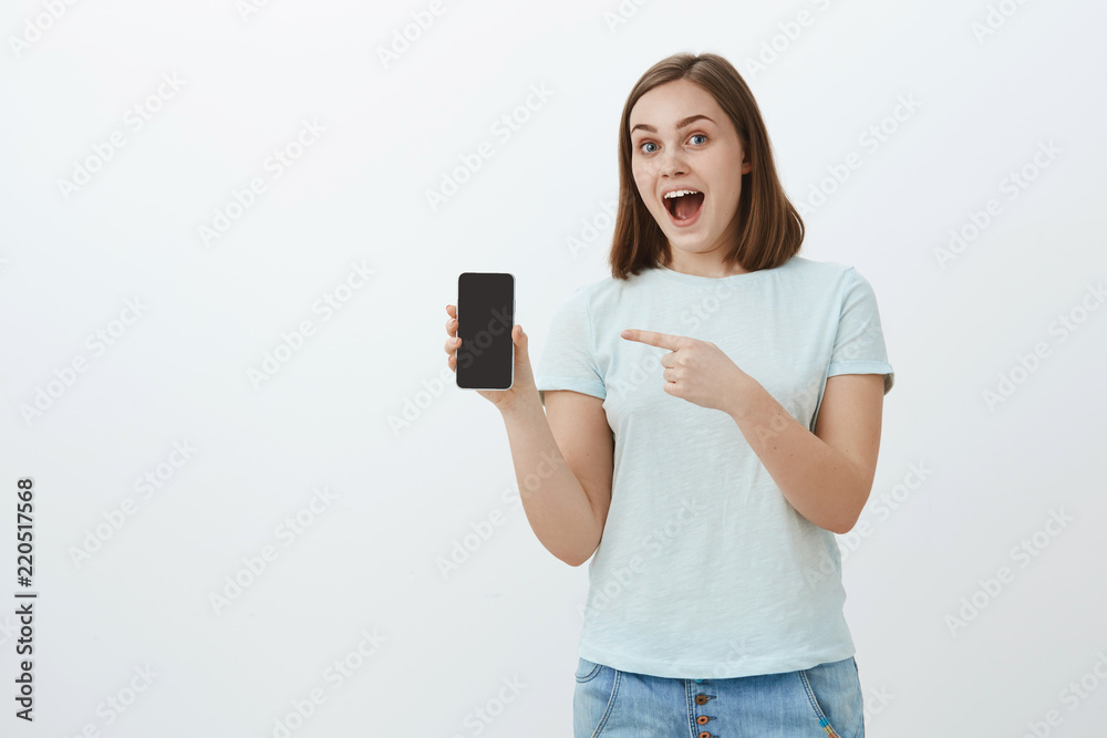 Fascinated good-looking young friendly female shop assistant in casual shirt showing smartphone screen and pointing at gadget talking about new features and cool design, smiling posing over grey wall Fascinated good-looking young friendly female shop assistant in casual shirt showing smartphone screen and pointing at gadget talking about new features and cool design, smiling posing over grey wall