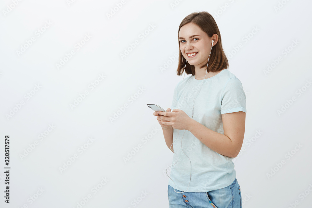 Never leave home without music. Portrait of charming friendly-looking delighted young woman in casual outfit getting ready walk to university wearing earphones and holding smartphone smiling aside Never leave home without music. Portrait of charming friendly-looking delighted young woman in casual outfit getting ready walk to university wearing earphones and holding smartphone smiling aside