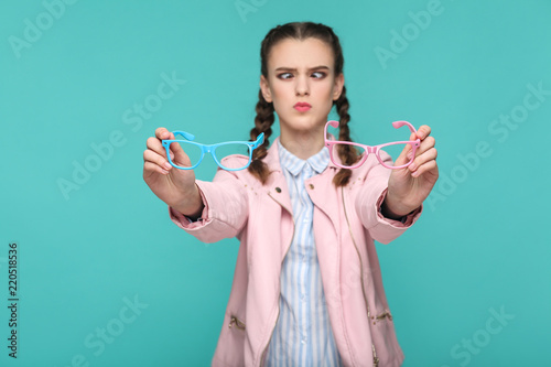 doubtful funny girl in casual or hipster style, pigtail hairstyle, standing, holding blue and pink glasses and looking at camera with crossed eye, Indoor studio shot, isolated on green background photo