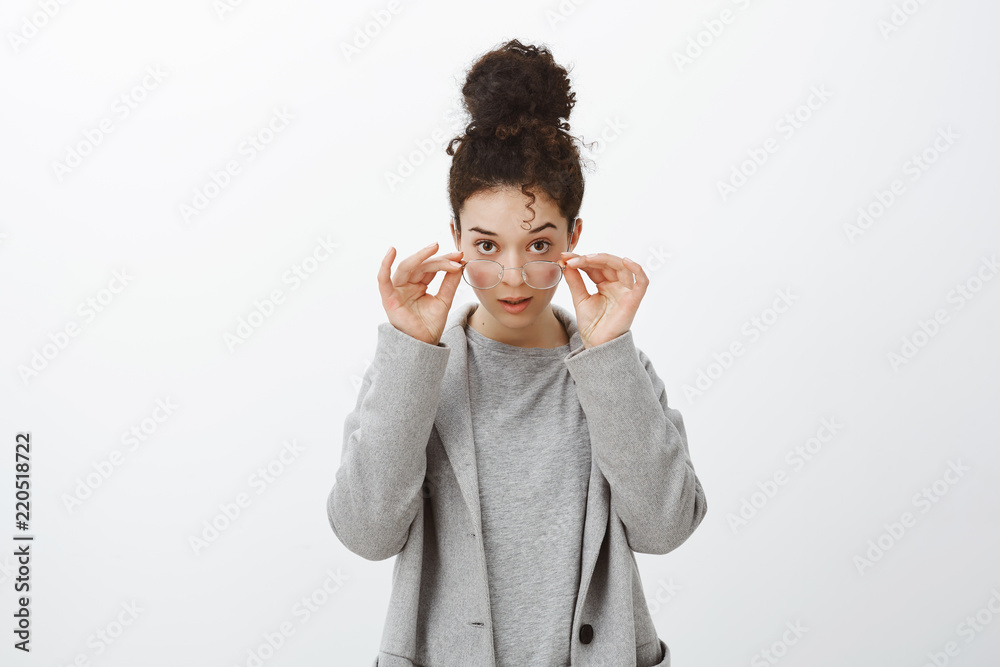Studio shot of shocked disbelieving attractive european girl with curly hair combed in bun, taking off glasses, looking from under forehead focused and intrigued, standing amazed over gray wall Studio shot of shocked disbelieving attractive european girl with curly hair combed in bun, taking off glasses, looking from under forehead focused and intrigued, standing amazed over gray wall