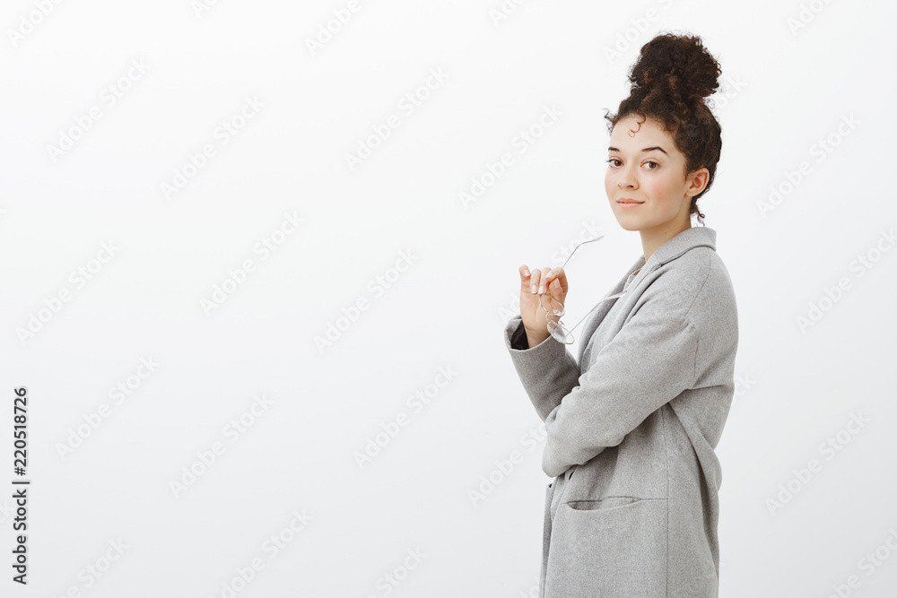 Portrait of confdent successful businesswoman in gray trendy coat, standing in profile and smiling self-assured at camera, holding glasses in hand, going in subway to work, standing against gray wall Portrait of confdent successful businesswoman in gray trendy coat, standing in profile and smiling self-assured at camera, holding glasses in hand, going in subway to work, standing against gray wall