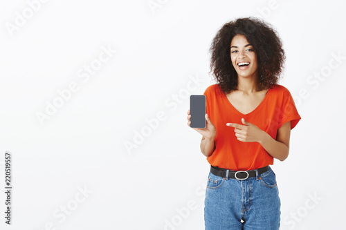 Best phone ever for silly price. Joyful confident and satisfied dark-skinned female holding smartphone, laughing and pointing at device with forefinger, advertising brand new gadget over grey wall photo