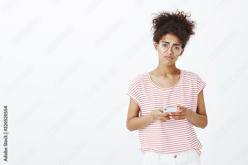 Girl feeling unhappy being dumbed via phone. Portrait of gloomy displeased cute woman in glasses and striped t-shirt, holding smartphone and frowning at camera, being offended with message Girl feeling unhappy being dumbed via phone. Portrait of gloomy displeased cute woman in glasses and striped t-shirt, holding smartphone and frowning at camera, being offended with message