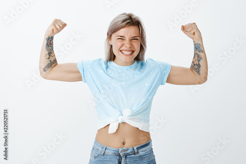 Show me real power of muscles. Portrait of good-looking playful and emotive girl with tattooed arms, raising hands and showing biceps while smiling broadly, working out for summer over grey wall