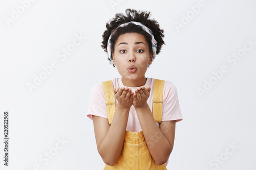 Sending warm kisses to my loving boyfriend. Cute and tender young woman with dark skin in trendy headband over hair and yellow overalls  folding lips and holding palms near mouth  giving mwah