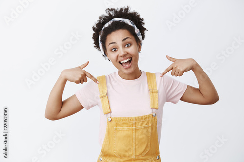 Good-looking charming active and optimistic African American girl in yeloow overalls and headband pointing at herself, tilting head and smiling broadly, being proud of her deeds over grey wall photo