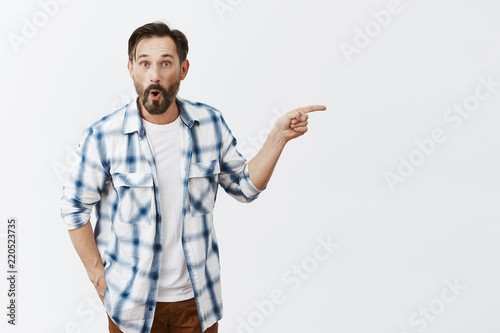 Guy impressed with car neighboor bought. Portrait of impressed and amazed handsome dad with beard in casual outfit, pointing right and gazing at camera with interest, asking question photo