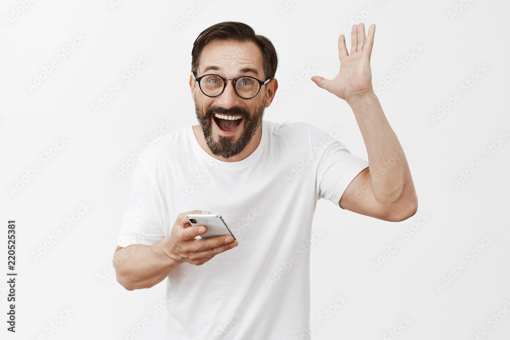 Yeah I did it. Portrait of excited happy and joyful grandad in glasses with beard, raising palm in joy and victory pose, holding smartphone and gazing with broad smile at camera, posing over grey wall Yeah I did it. Portrait of excited happy and joyful grandad in glasses with beard, raising palm in joy and victory pose, holding smartphone and gazing with broad smile at camera, posing over grey wall