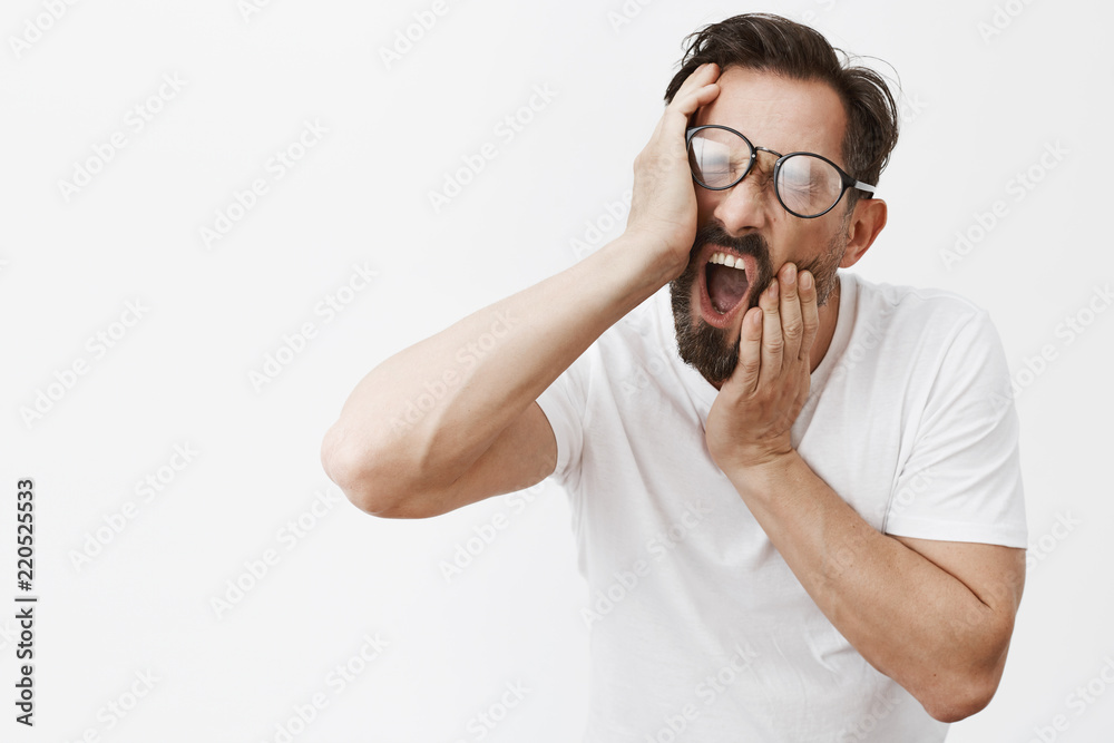 Man cannot wake up, trying pull his face to cheer up. Portrait of funny caucasian bearded male with dark hair, squeezing cheeks and yawning, being tired and sleepy, wanting go to bed over grey wall Man cannot wake up, trying pull his face to cheer up. Portrait of funny caucasian bearded male with dark hair, squeezing cheeks and yawning, being tired and sleepy, wanting go to bed over grey wall