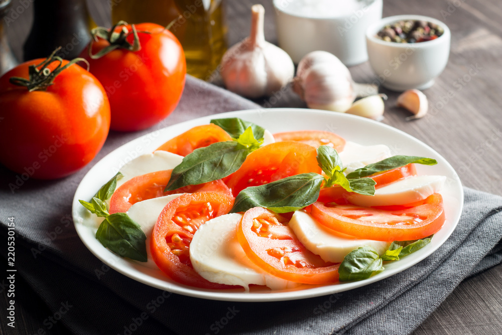 Photo of Caprese Salad with tomatoes, basil, mozzarella, olives and olive oil on wooden background. Italian traditional caprese salad ingredients. Mediterranean, organic and natural food concept.