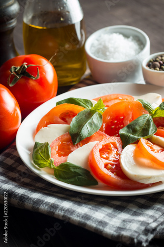 Photo of Caprese Salad with tomatoes, basil, mozzarella, olives and olive oil on wooden background. Italian traditional caprese salad ingredients. Mediterranean, organic and natural food concept.