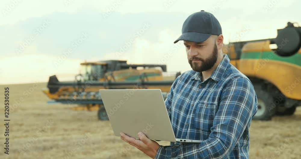 Portrait shot of the Caucasian middle aged farmer working in the field on the laptop computer on the combine machinery background. Portrait shot of the Caucasian middle aged farmer working in the field on the laptop computer on the combine machinery background.