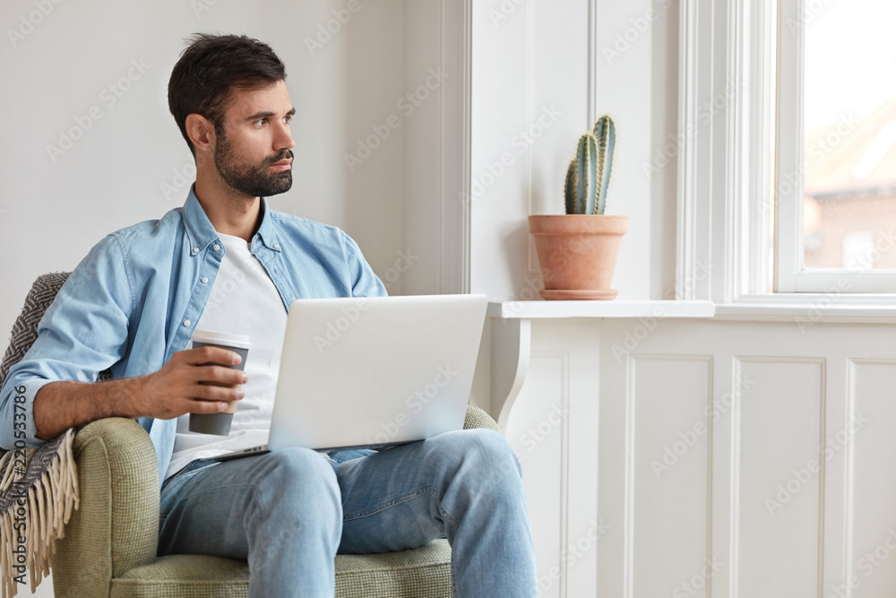 Pensive bearded blogger, sits in armchair in cozy room, works on laptop computer, drinks takeaway coffee, ponders on idea for publication, poses over home interior. Entrepreneur works on startup