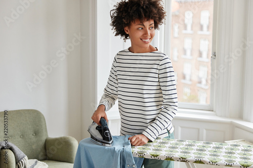 Photo of cheerful black housewife does housework at home during weekend, irons clothes on ironing board, looks joyfully aside. Lovely dark skinned wife irons husbands shirt who is going on work photo