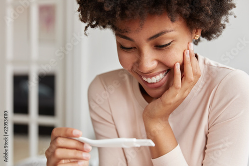 Close up shot of happy future mother smiles broadly when finds out positive results of pregnancy test, cant believe in such good news, poses against cozy domestic background. Yes, I am pregnant!