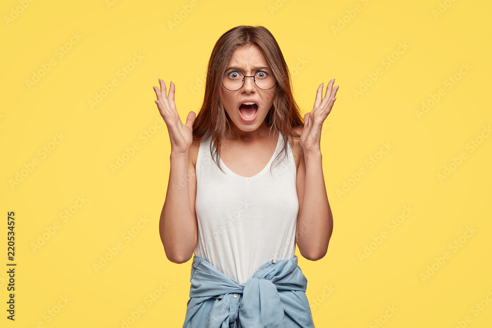 Outraged angry emotional woman being at loss, looks perplexed and furious, gestures actively while quarrels and defends her opinion, wears denim shirt around waist, stands against yellow wall Outraged angry emotional woman being at loss, looks perplexed and furious, gestures actively while quarrels and defends her opinion, wears denim shirt around waist, stands against yellow wall