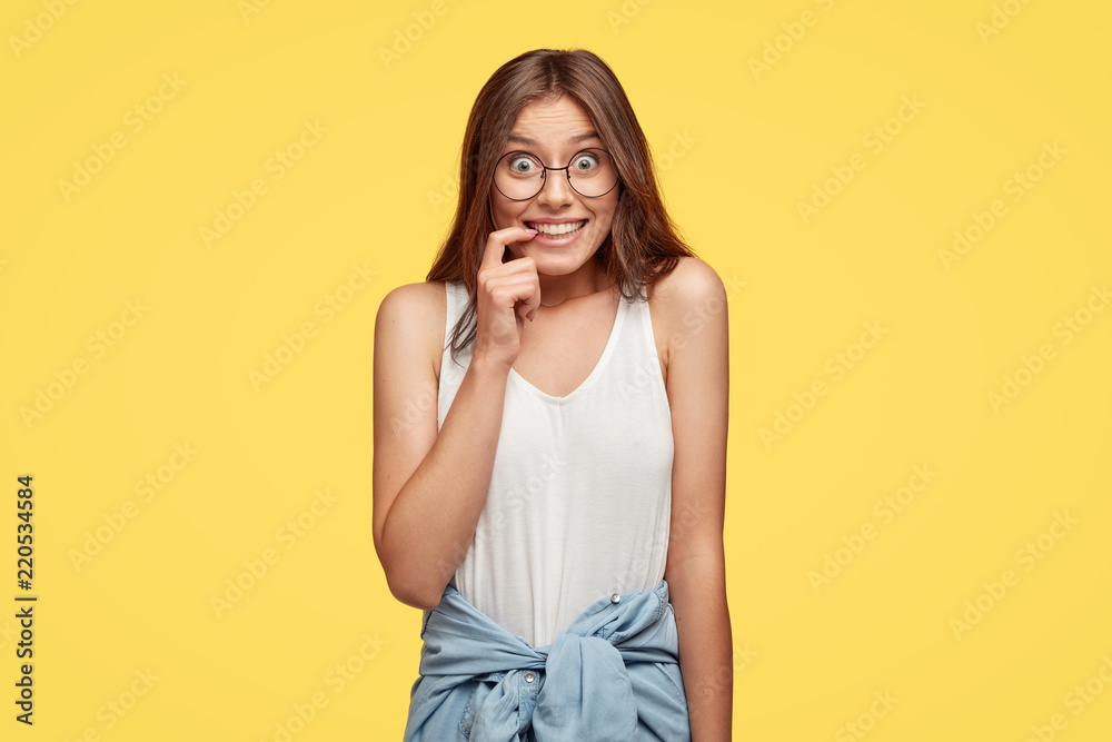 Optimistic satisfied teenager has happy expression, keeps fore finger near mouth, looks positively at camera, happy to meet with coevals, stands against yellow background. People and emotions concept Optimistic satisfied teenager has happy expression, keeps fore finger near mouth, looks positively at camera, happy to meet with coevals, stands against yellow background. People and emotions concept