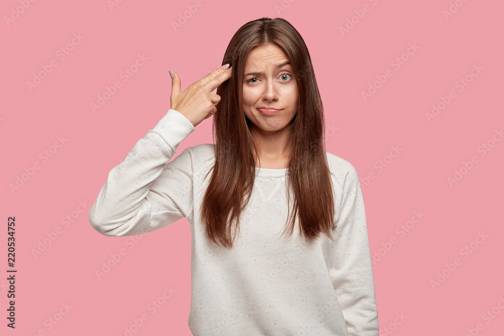 You are killing me! Exhausted student makes suicide gesture, shoots in temple, commits suicide, wears casual outfit, stands against pink background. Female feels bored, looks displeased at camera You are killing me! Exhausted student makes suicide gesture, shoots in temple, commits suicide, wears casual outfit, stands against pink background. Female feels bored, looks displeased at camera