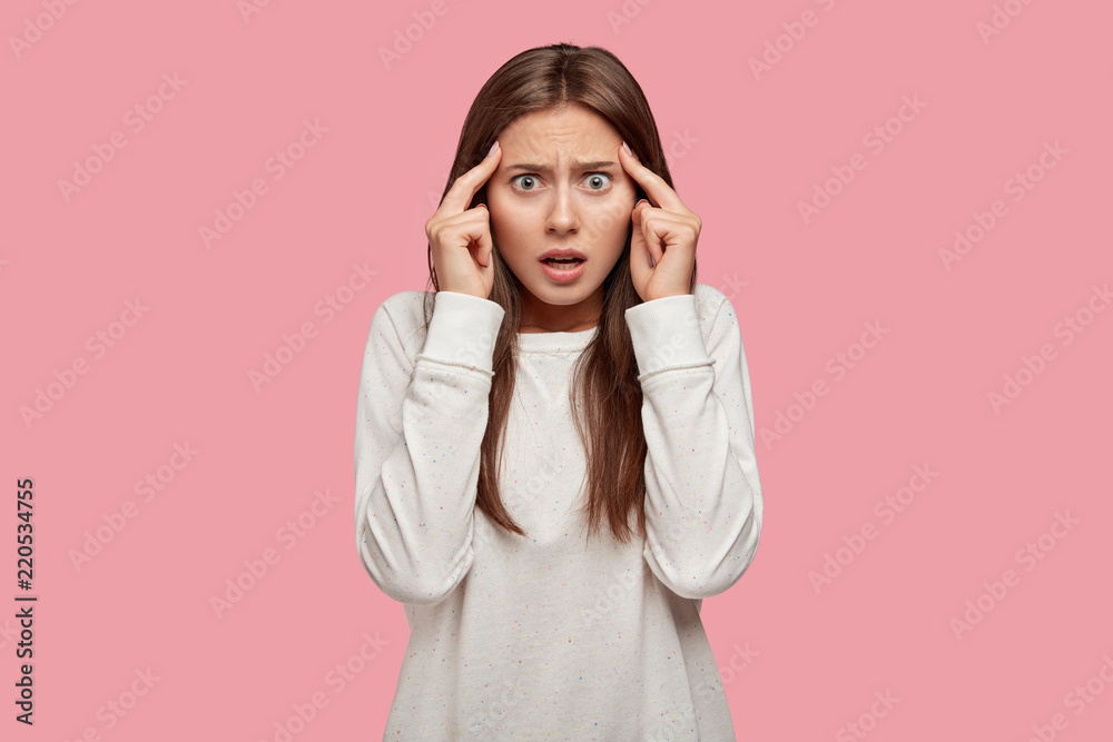Photo of displeased student stares with bugged eyes, keeps fingers on temples, has stressful situation at university, stands against pink background. Young woman afraids something, recollects in mind Photo of displeased student stares with bugged eyes, keeps fingers on temples, has stressful situation at university, stands against pink background. Young woman afraids something, recollects in mind