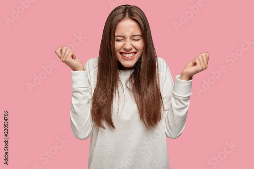Successful brunette girl closes eyes from happiness, keeps raised fists, dressed casually, celebrates her victory with triumph, isolated over pink background. People, joy, body language concept photo