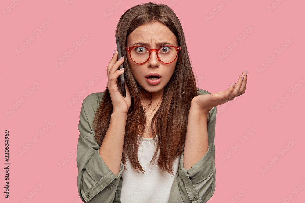 Angy female model has telephone conversation, looks surprisingly at camera, gestures with hand, looks with puzzled expression, stands against pink wall. Emotive woman talks unpleasantly indoor