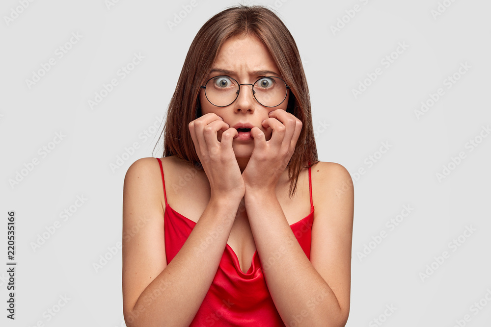 Worried brunette young woman looks with anxious expression, keeps hands near mouth, has dark hair, dressed in casual domestic clothes, round transparent glasses, isolated over white background Worried brunette young woman looks with anxious expression, keeps hands near mouth, has dark hair, dressed in casual domestic clothes, round transparent glasses, isolated over white background
