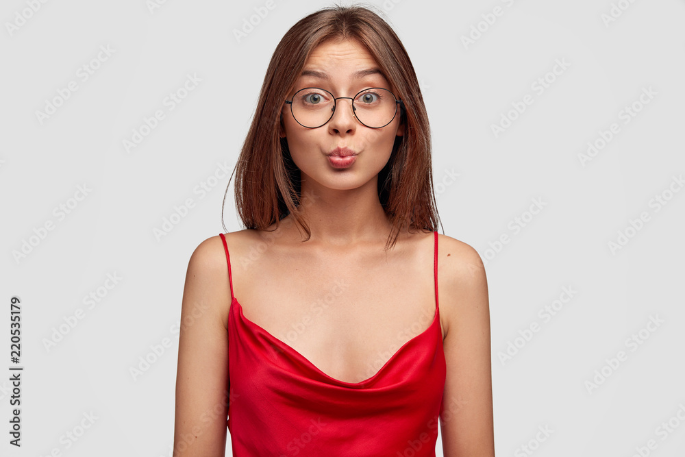 Cute girlfriend looks coquette, rounds lips, sends kisses at camera, wears round glasses and red stylish nightdress, has healthy skin, stands over white background. Human facial expressions concept Cute girlfriend looks coquette, rounds lips, sends kisses at camera, wears round glasses and red stylish nightdress, has healthy skin, stands over white background. Human facial expressions concept
